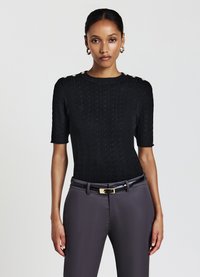 Schwarzes gestricktes Kurzarmoberteil mit Zopfmuster und goldenen Knopfdetails an den Schultern, kombiniert mit grauen maßgeschneiderten Hosen und einem schwarzen Gürtel.