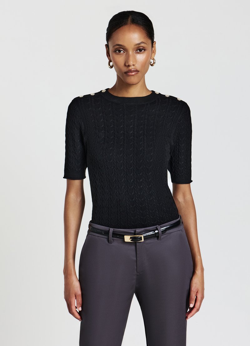 Schwarzes gestricktes Kurzarmoberteil mit Zopfmuster und goldenen Knopfdetails an den Schultern, kombiniert mit grauen maßgeschneiderten Hosen und einem schwarzen Gürtel.