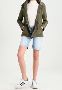 Chaqueta verde con capucha, camiseta beige, pantalones cortos de mezclilla claros y zapatillas blancas con rayas negras. Enfocada en un diseño práctico y un estilo casual.