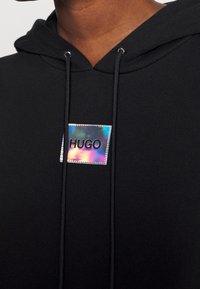 Svart hoodie med en mjuk textur som har en holografisk etikett märkt "HUGO" och silverdetaljer på snörningen.