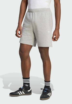 Osoba nosząca jasnoszorty Adidas, czarne skarpety Adidas z białymi paskami oraz czarne buty sportowe Adidas z białymi paskami.
