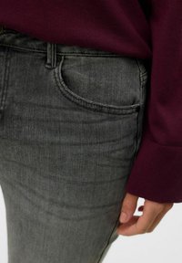 I jeans in denim grigio presentano un classico design a cinque tasche con cuciture visibili. La vita è accentuata dal polsino di un maglione borgogna.