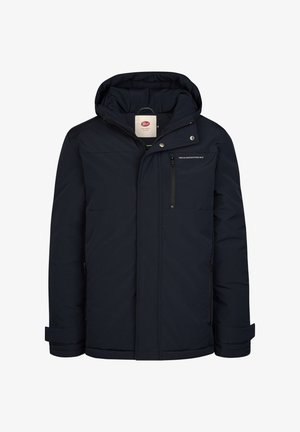 Rødhåret dreng står iført en marineblå hættetrøje, blå sweatshirt, revnede lyseblå jeans og hvide sneakers mod en ensfarvet baggrund.