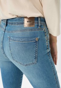 Primo piano di una persona che indossa jeans azzurri chiari con borchie metalliche sui lati e un'etichetta in pelle marrone con la scritta "Brax" sulla vita.