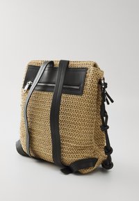 Vevd stråryggsekk med svarte skinnakenter, justerbare stropper og en glidelåslomme foran. Teksturert, strukturert form og slitesterkt materiale.