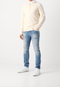 Sweter w kolorze kremowym z kołnierzem na pół-zamek, w połączeniu z jasnoniebieskimi dżinsami. Białe tenisówki dopełniają stylizację. Ogólna gładka tekstura.