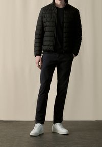 Czarna kurtka puchowa z wysokim kołnierzem, zestawiona z czarną bluzką, granatowymi spodniami i białymi sneakersami. Gładka tekstura i prosty design.