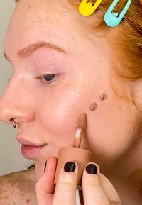 Flytande highlighter i en brun behållare med applikator med mjuk spets, applicerad på hud med fräknar. Modellen har röd hårfärg och svarta naglar.