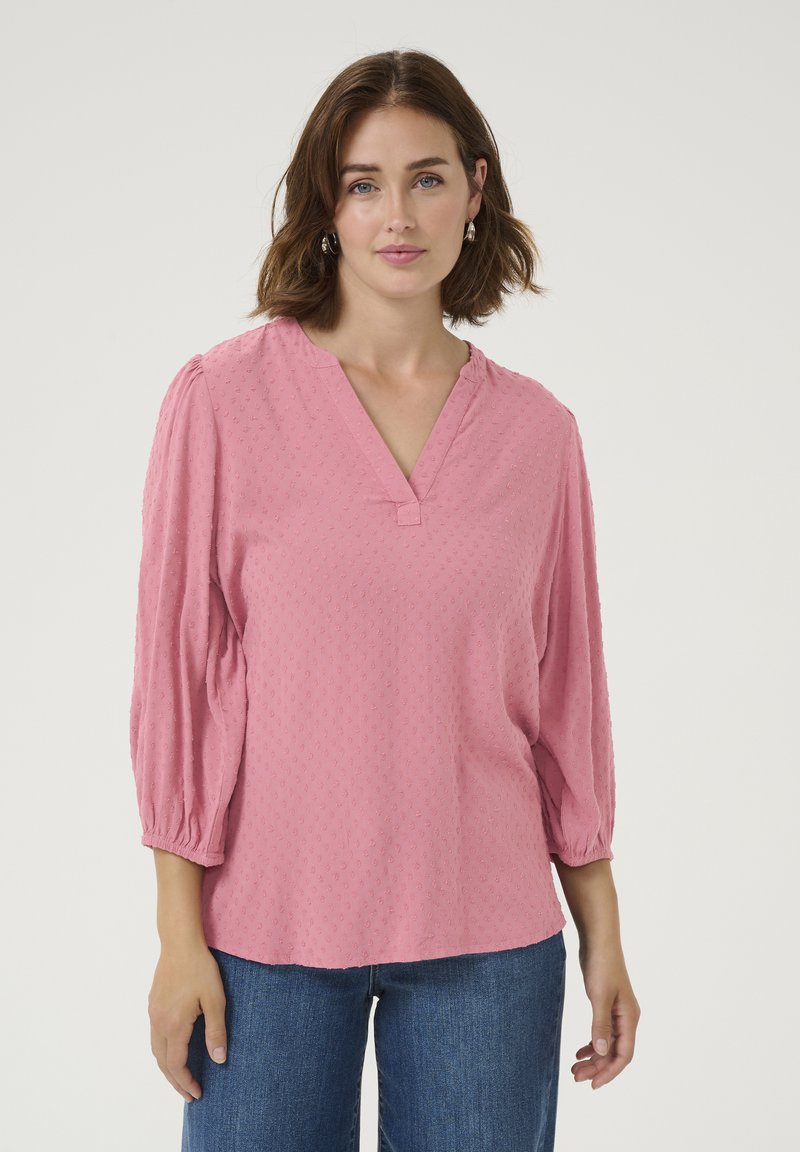Blouse rose, texturée, avec un décolleté en V et des manches bouffantes trois-quarts. Le tissu présente un motif subtil et une coupe décontractée.