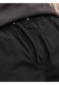 Svarta shorts med elastisk midja, som har en dragsnöre och sammanknuten textur. Bakgrunden visar ett mörkgrått tyg som överlappar midjan.