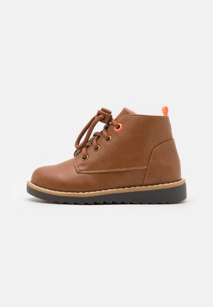 Brun læder ankelstøvle med rund tå, orange snøreløbere, sort gummisål og justerbare snørebånd med vippefunktion mod en hvid baggrund.