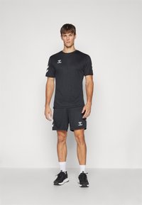 Conjunto deportivo negro que incluye una camiseta de manga corta y pantalones cortos. Ajuste atlético, textura suave, logotipo blanco y detalles blancos en las mangas.