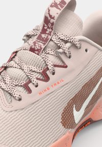 Chaussure de trail Nike beige et rose avec lacets à motifs, tissu texturé et virgule blanche Nike sur le côté.