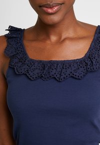 Mørkeblå tanktop med en bølget halsudskæring med øjelet-detaljer, ribbet tekstur og brede skulderstropper.