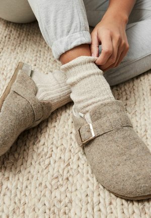 Sabots gris en feutre avec une boucle, associés à des chaussettes côtelées crème, sur un tapis beige texturé. Des matériaux doux et confortables sont évidents.