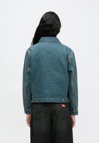 Άτομο με μακριά μαύρα μαλλιά που φοράει μπλε τζιν μπουφάν και μαύρο τζιν παντελόνι, στέκεται με την πλάτη προς τα εμπρός σε απλό φόντο.