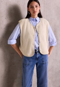 Gilet en polaire beige avec des boutons-pression, ourlet arrondi, porté par-dessus une chemise à rayures bleu clair. Le modèle pose avec les mains sur les hanches, portant un pantalon en denim décontracté.