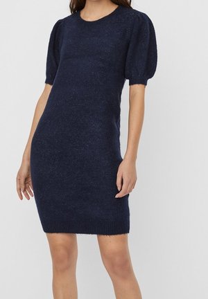 Frau trägt ein marineblaues Strickkleid mit kurzen Puffärmeln und einem gerippten Saum, steht vor einem schlichten hellen Hintergrund.