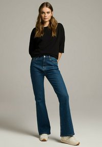 Rozszerzane niebieskie dżinsy z wysokim stanem, wykonane z materiału dżinsowego. Połączone z czarną bluzką z długim rękawem i białymi trampkami. Gładka tekstura.