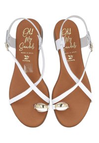 Sandalias blancas con tiras y suela plana, que cuentan con un acento dorado en la punta, una plantilla marrón y correas ajustables en el tobillo. Materiales ecológicos utilizados.