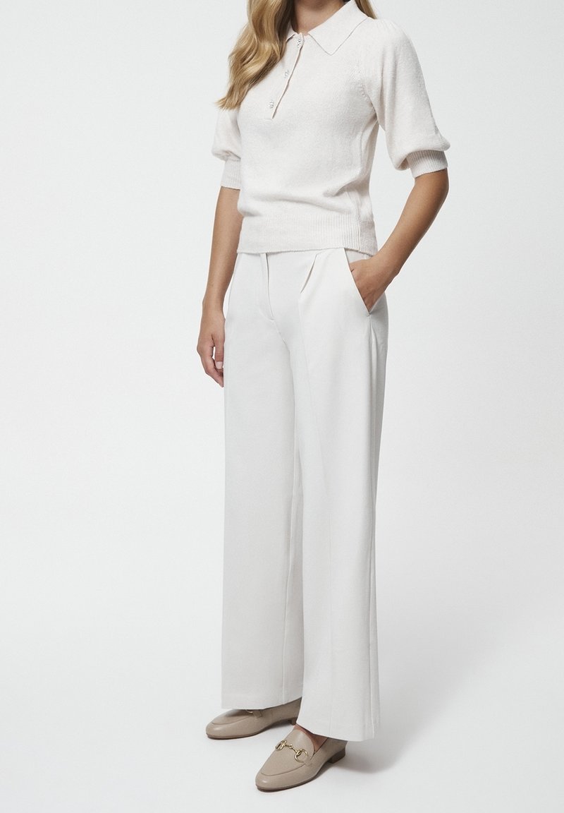 Femme portant un pull en maille blanc à manches courtes bouffantes, un pantalon blanc à jambes larges et des mocassins beiges avec un détail doré, debout avec une main dans la poche.