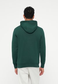 Tamsiai žalia hoodie su minkšta tekstūra, turi erdvų kapišoną, gumbines rankoves ir apatinius kraštus. Gale matomas paprastas dizainas be logotipų.