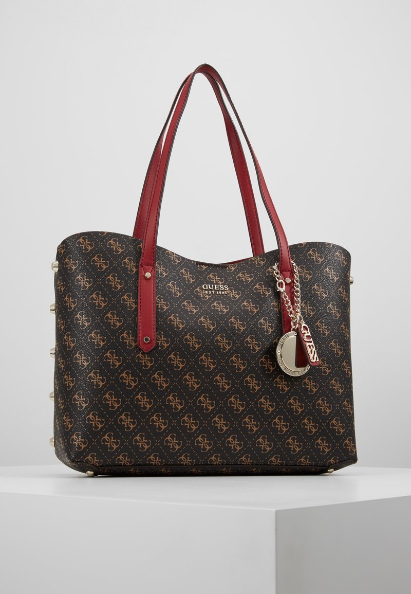 Bruine monogram handtas met rode synthetische handvatten, gouden hardware accenten en decoratieve studs aan één zijde. Merklogo duidelijk zichtbaar.