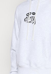 Hoodie gris clair en mélange de coton, avec une poche kangourou et une impression graphique noire sur la poitrine, comprenant des yeux et des formes abstraites.
