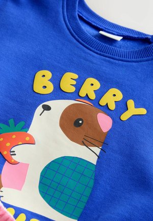 Kék póló, amelyen egy tengerimalac látható, amint egy epret tart, és az állat felett nagy sárga betűkkel a "BERRY" felirat szerepel.