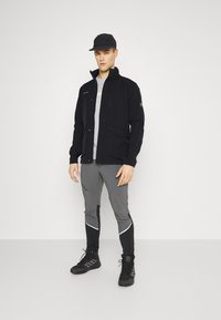 Veste noire avec zip et poches à rabat, assortie à un pantalon gris avec des accents noirs. Le modèle porte une casquette noire et des bottes. Texture lisse.