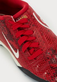 Röd sneaker med ett texturerat skinnmönster, vit logotyp, platta röda snören och svart gummisula. Har en vadderad röd krage.
