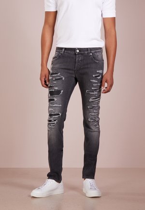 Muž v černých roztrhaných úzkých džínách, bílých teniskách a jednoduchém bílém tričku s krátkým rukávem stojící na béžové podlaze.