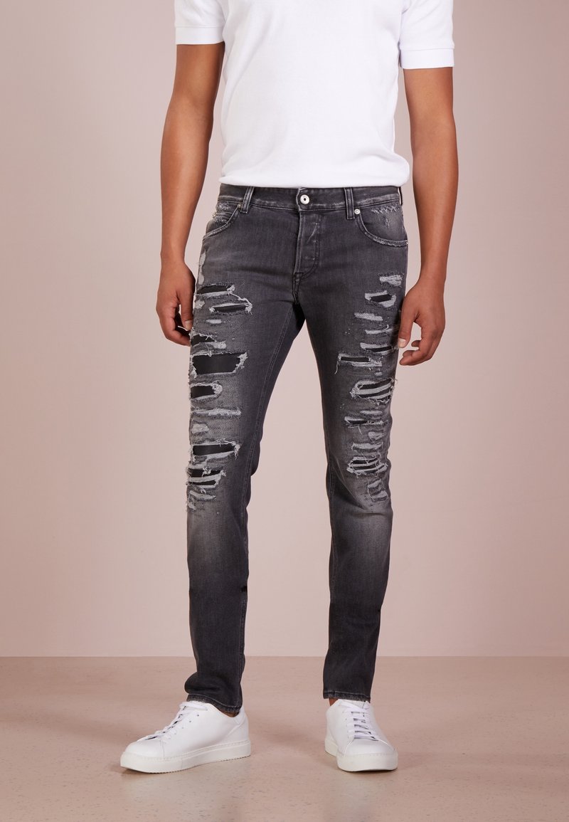 Homme portant un jean skinny noir déchiré, des baskets blanches et une chemise blanche à manches courtes unie, debout sur un sol beige.