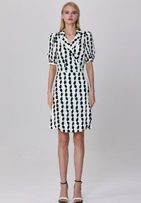 Robe blanche courte avec un motif circulaire vert et noir, dotée d'un col et de manches bouffantes ; tissu lisse, coupe au genou. Sandales à talons noires.