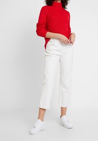 Pull rouge en maille avec un col montant, associé à un jean blanc raccourci aux ourlets effilochés, et des baskets blanches.