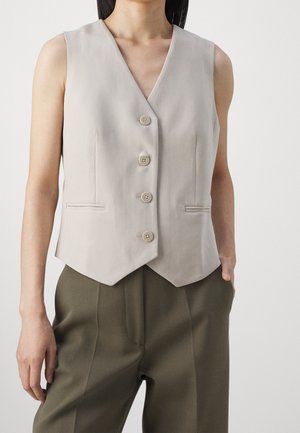 Beżowa kamizelka bez rękawów na guziki z dekoltem w serek, noszona na oliwkowozielonych spodniach, ręce swobodnie opuszczone wzdłuż ciała, neutralne tło.