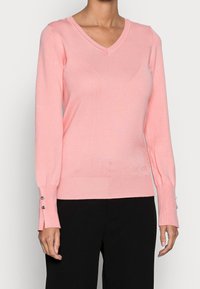 Femme portant un pull V en rose clair avec des manches longues et un pantalon noir, le pull a de petits boutons aux poignets et un logo discret près de l'ourlet.