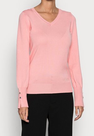 Frau trägt einen hellrosa V-Ausschnitt-Pullover mit langen Ärmeln und schwarze Hosen. Der Pullover hat kleine Knöpfe an den Ärmelbündchen und ein dezentes Logo am Saum.