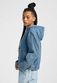 Vaaleansininen farkkudekotinko, jossa on kengurupussitasku, nauhalla säädettävä huppu ja rento leikkaus. Takissa on näkyvät saumat ja ribbireunus.