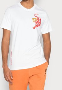 Vit bomullströja med en röd tiger grafik på bröstet, tillsammans med en orange Nike-logotagg och vertikal röd text på sidan.