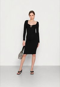 Vestido preto de manga longa com silhueta ajustada, apresentando detalhes franzidos e um laço no decote. Combinado com uma mala riscada e saltos pretos.