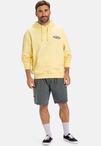 Yellow hoodie with front pocket and drawstrings, featuring a logo in black and pink. Paired with gray shorts and black sneakers.