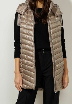 Femme portant une veste matelassée beige sans manches avec une capuche sur un haut noir à manches longues et un pantalon noir, debout devant un fond uni.
