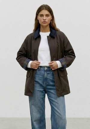Femme portant une veste sombre avec des poignets et un col à carreaux par-dessus une chemise blanche et un jean bleu, debout devant un fond clair uni.