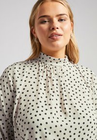 Blusa crema con pois neri, colletto alto, dettaglio arricciato al collo e tessuto morbido e fluido.