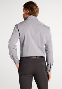Graues Langarmhemd mit Button-Down-Kragen, kombiniert mit dunklen Hosen mit glatter Textur und zwei Gesäßtaschen.