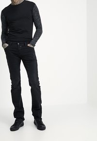 Svart långärmad skjorta med mörkgrå texturerade ärmar, kombinerad med svarta jeans och svarta sneakers, stående framför en helt vit bakgrund.