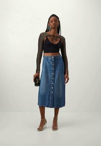 Junge Frau trägt ein schwarzes Crop-Top mit langen Ärmeln, einen knöpfbaren blauen Denim-Midikleid, transparente Riemchen-Absätze und hält eine kleine schwarze Clutch.