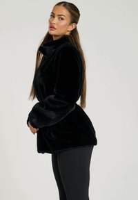 Giacca in pelliccia sintetica nera con colletto alto, maniche lunghe e vita con cintura, caratterizzata da una texture morbida e una vestibilità rilassata. Indossata con leggings neri.