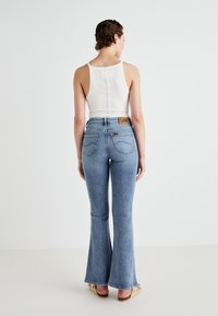 Lichtblauwe flared jeans met een hoge taille, voorzien van een bruine leren label op de achterkant. De top is een witte tanktop met knoopdetails op de rug.